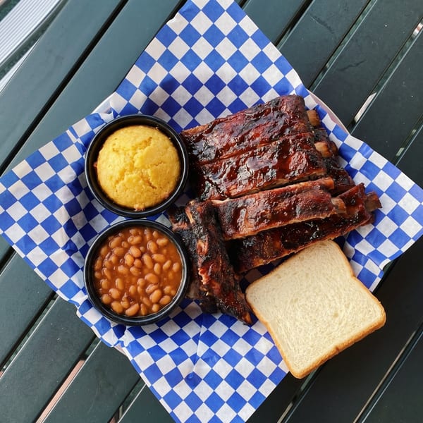 Barbecue ribs with baked beans, cornbread, and white bread on blue and white checkered paper.