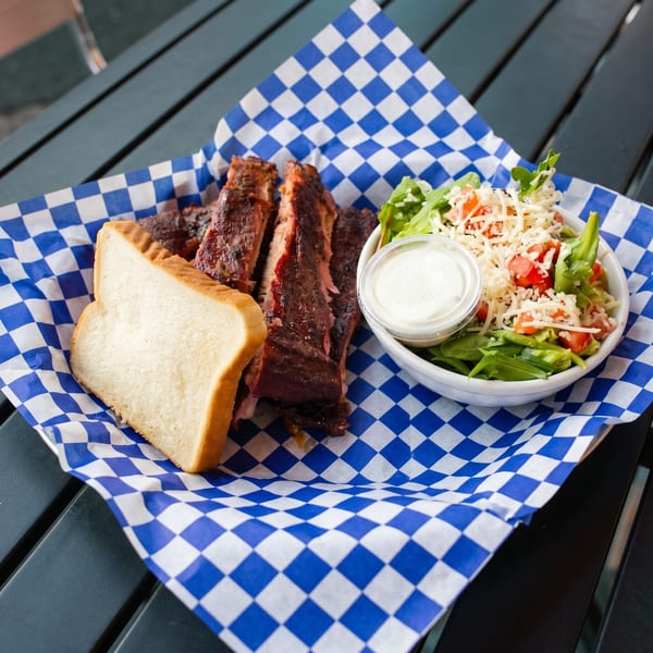 BBQ ribs, a slice of white bread, and a side salad with dressing on blue and white checkered paper.