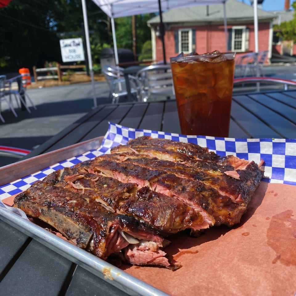 Sliced BBQ ribs on a tray with checkered paper, iced tea, and a red building in the background.