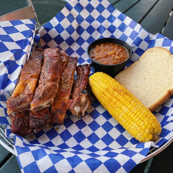 BBQ ribs, baked beans, corn on the cob, and white bread in a blue and white checkered basket.