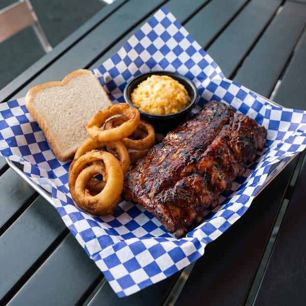 BBQ ribs, onion rings, mac and cheese, and white bread on a blue and white checkered paper.