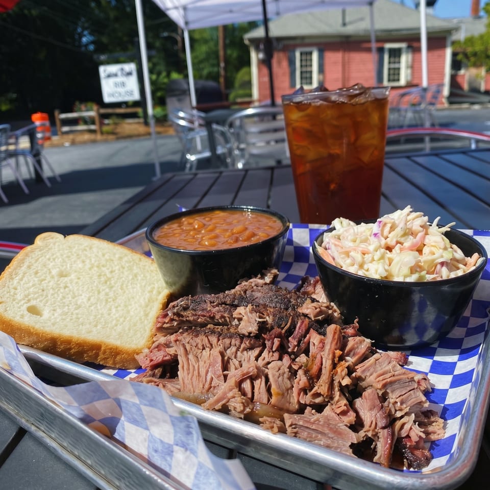 A BBQ platter with pulled pork, baked beans, coleslaw, and white bread on an outdoor table.